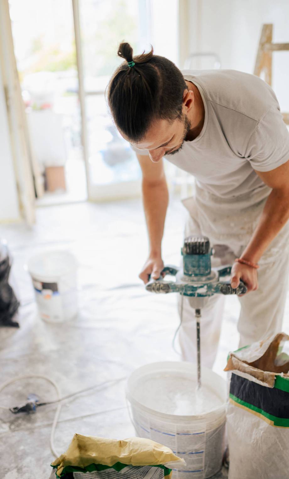 A plasterer is preparing material in a bucket for plastering while standing in house in a renovation process. A manual worker is standing in an apartment and preparing plaster for skim coating.