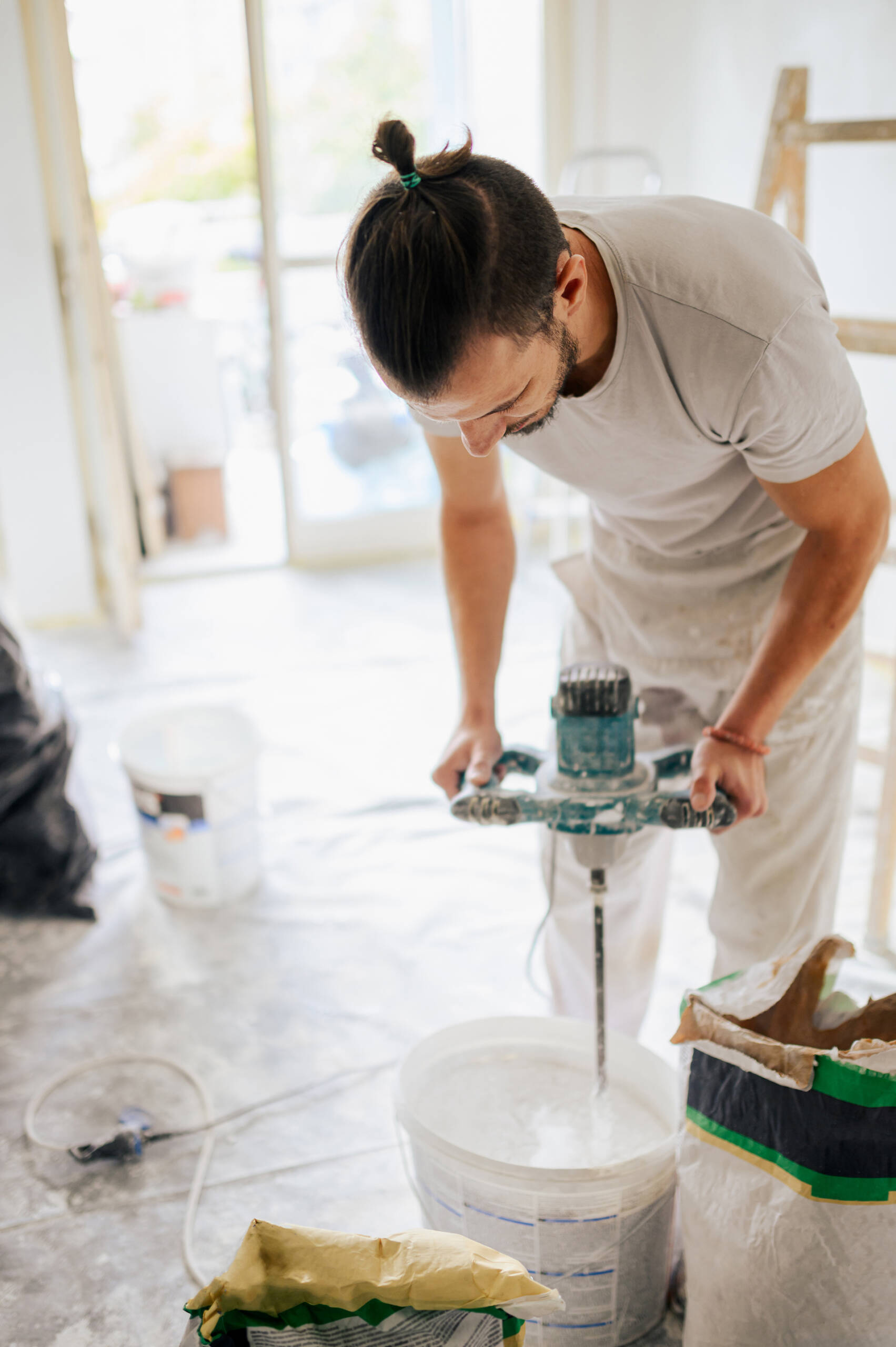 A plasterer is preparing material in a bucket for plastering while standing in house in a renovation process. A manual worker is standing in an apartment and preparing plaster for skim coating.